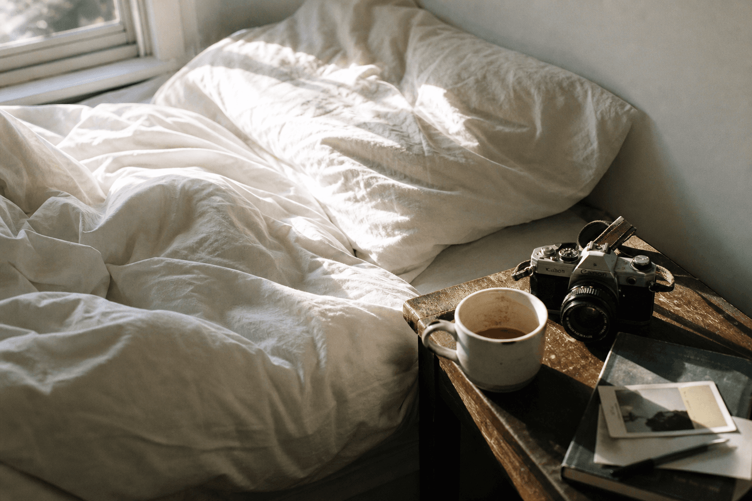 Morning light on an unmade bed with rumpled linen and a worn film camera on the nightstand